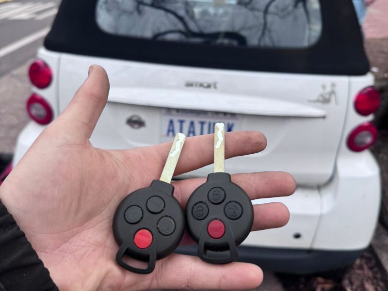 A hand holding two car keys in front of a parked Smart car. Car Key Replacement: A hand holding two car keys in front of a parked Smart car.